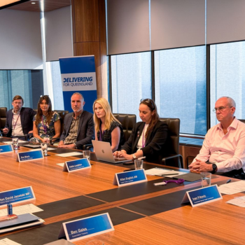 Six people seated around a boardroom table with name plaques, in front of a "Delivering for Queensland" banner and city-view windows.